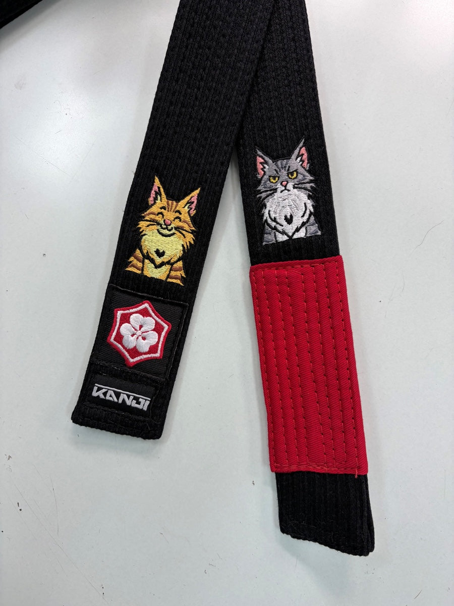Two real black belt ends with yellow and grey cat embroidery, the original KANJI labels on one end and a red rank bar on the other.